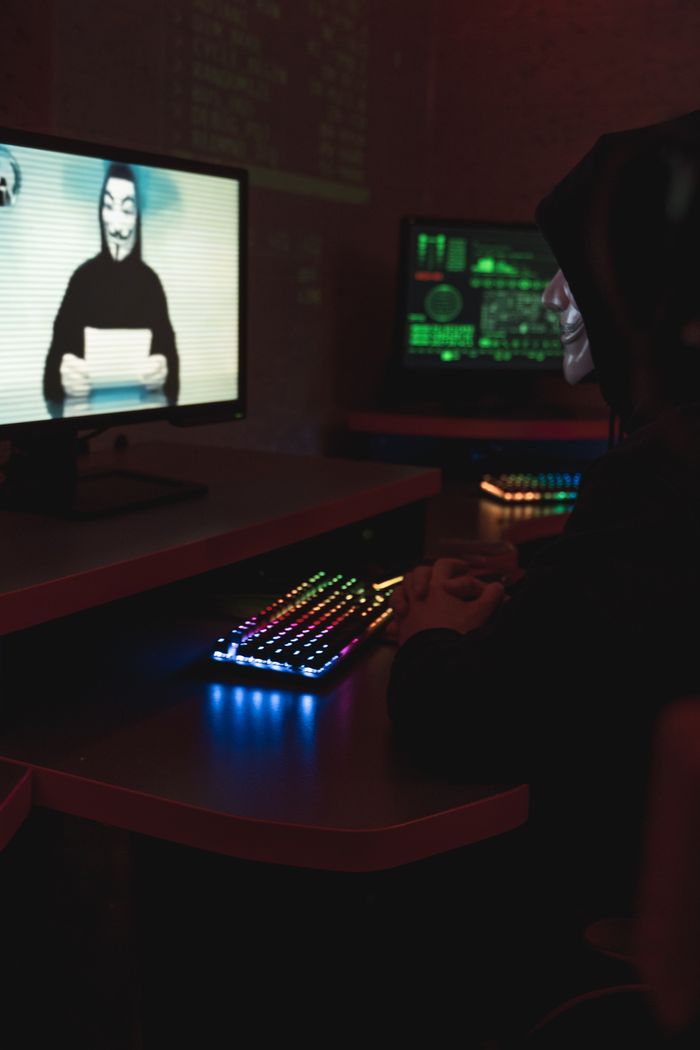 A hacker wearing a Guy Fawkes mask sitting in a dimly lit room with computers displaying code.